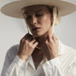 A person in a white shirt wears the Palmer Hat by Janessa Leone and holds organic cotton ties with regenerative leather aglets, eyes closed, against a neutral background.