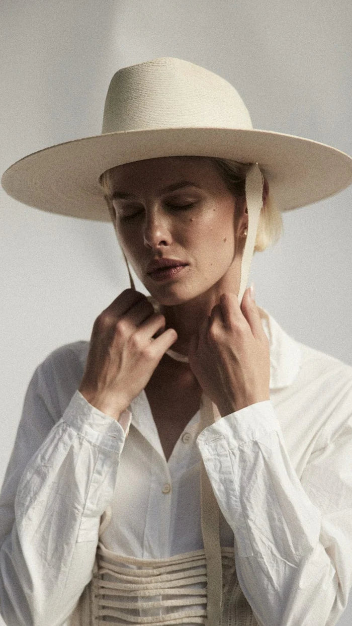 A person in a white shirt wears the Palmer Hat by Janessa Leone and holds organic cotton ties with regenerative leather aglets, eyes closed, against a neutral background.