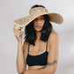 A person wearing the elegant Margot Visor by Gigi Burris Millinery and a black top against a plain background, seamlessly combining sophistication with the practicality of crochet raffia style.