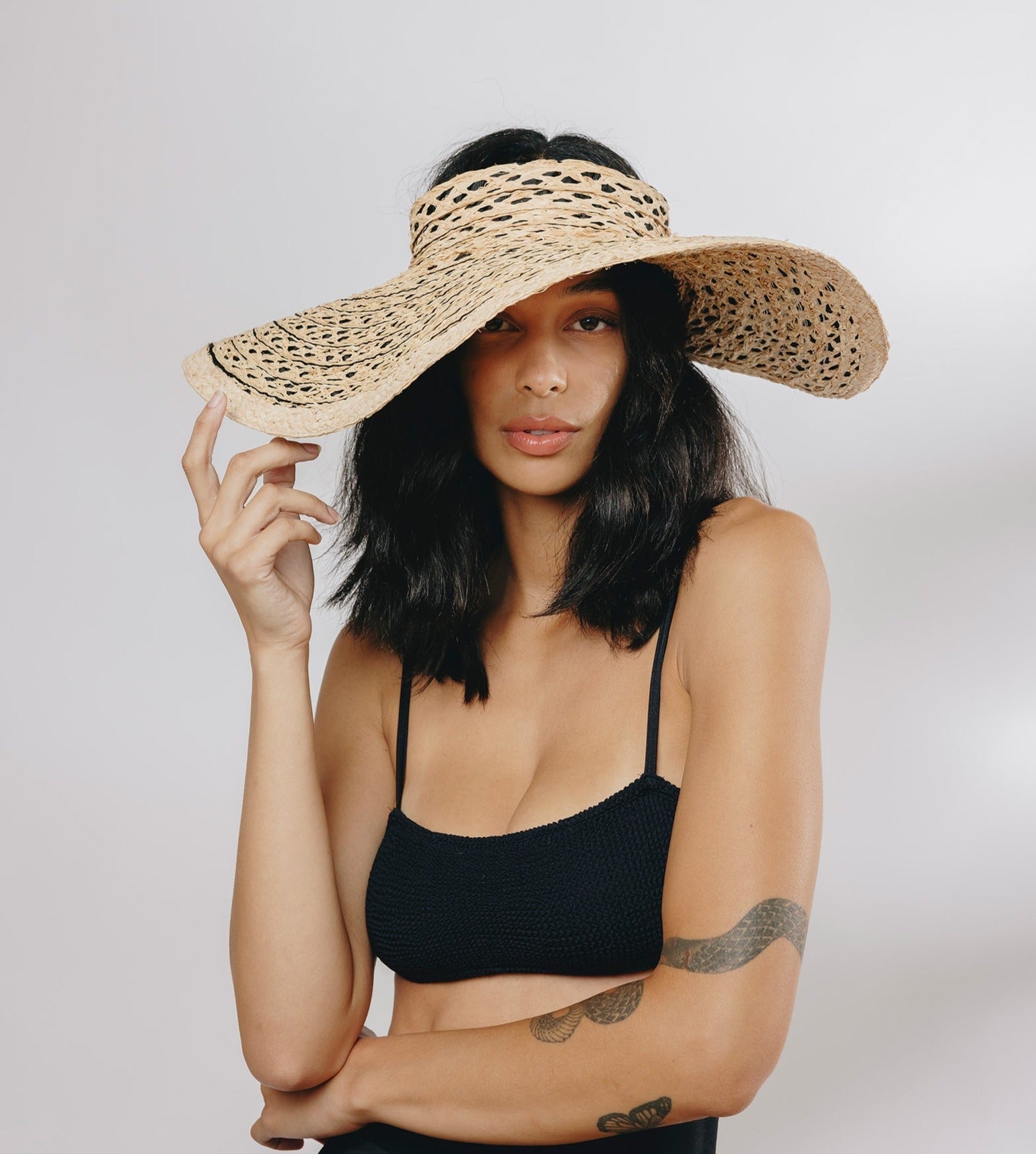 A woman wears a black top and the Gigi Burris Margot Visor, elegantly showcasing this signature piece from Gigi Burris, as she holds the brim with one hand against a plain background.