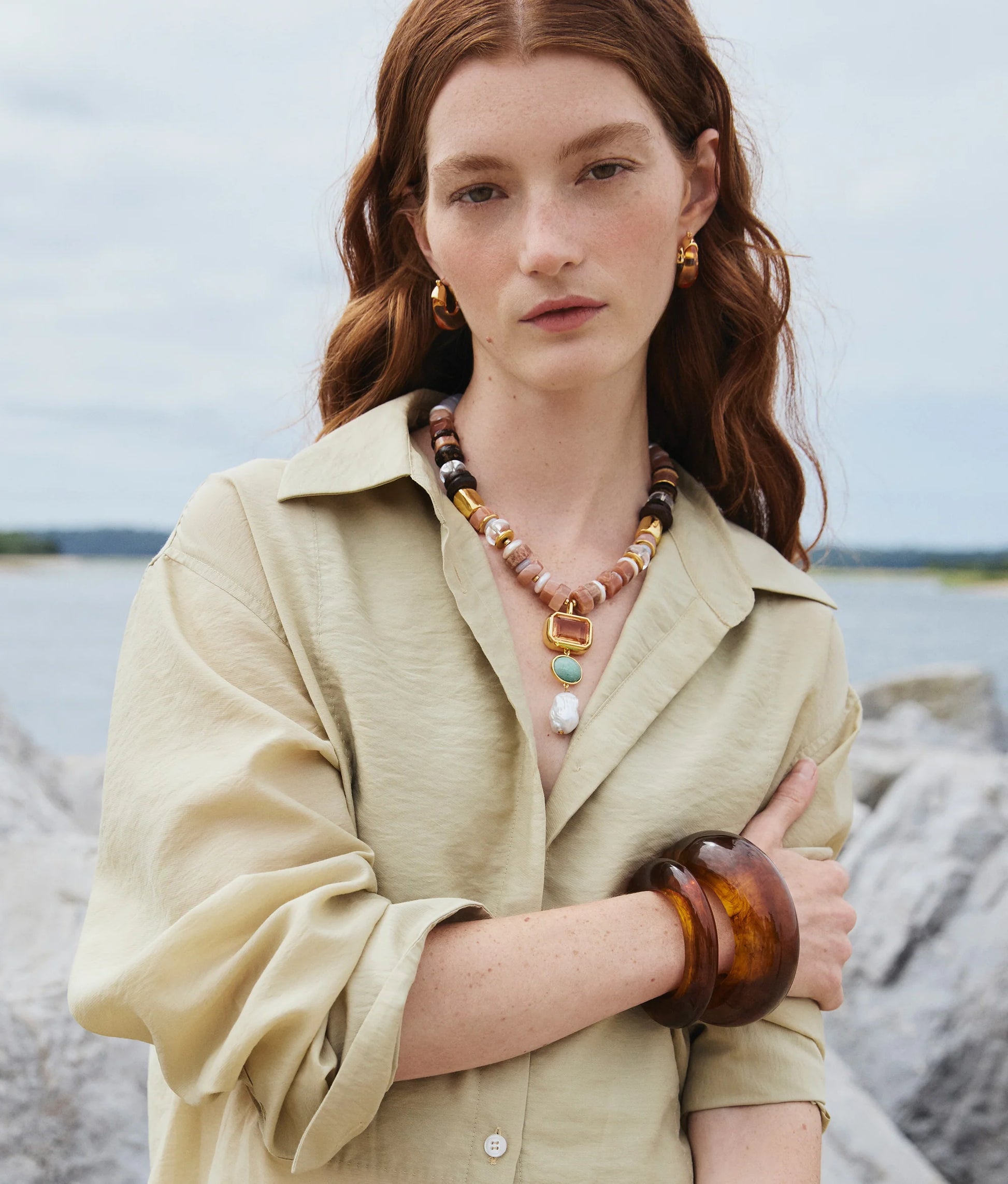A woman with long red hair stands outdoors near water and rocks, wearing a beige shirt and the Lizzie Fortunato Ridge Cuff—chunky gold and brown jewelry.