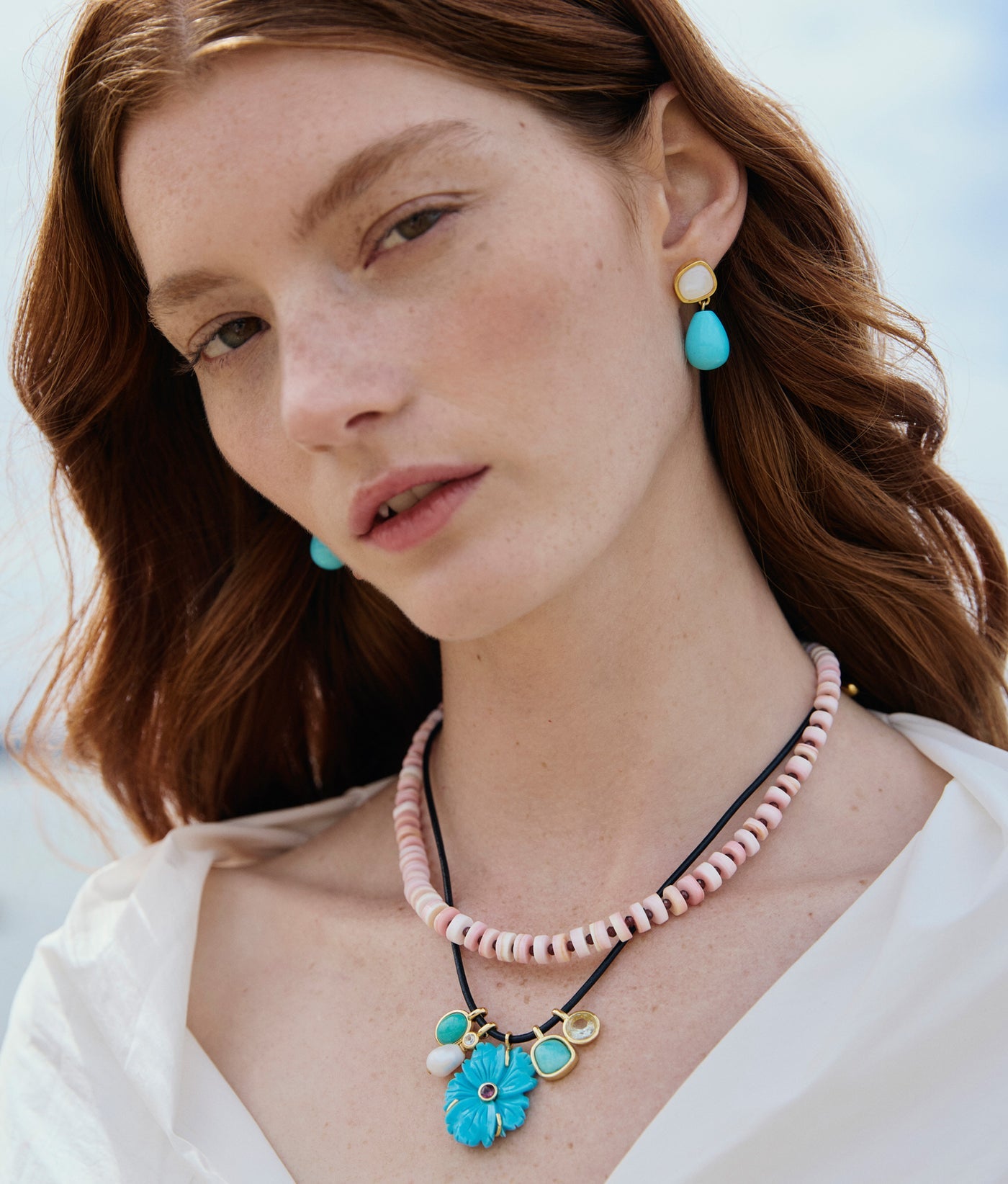 A woman with fair skin and long red hair wears a white blouse, turquoise bead drops, and Lizzie Fortunato's Rock Pool Earrings, paired with layered necklaces featuring pink beads and a turquoise flower pendant.