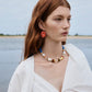 A woman with long red hair stands outdoors near the water, wearing a white blouse, a colorful beaded necklace, and Lizzie Fortunato’s New Bloom Earrings.