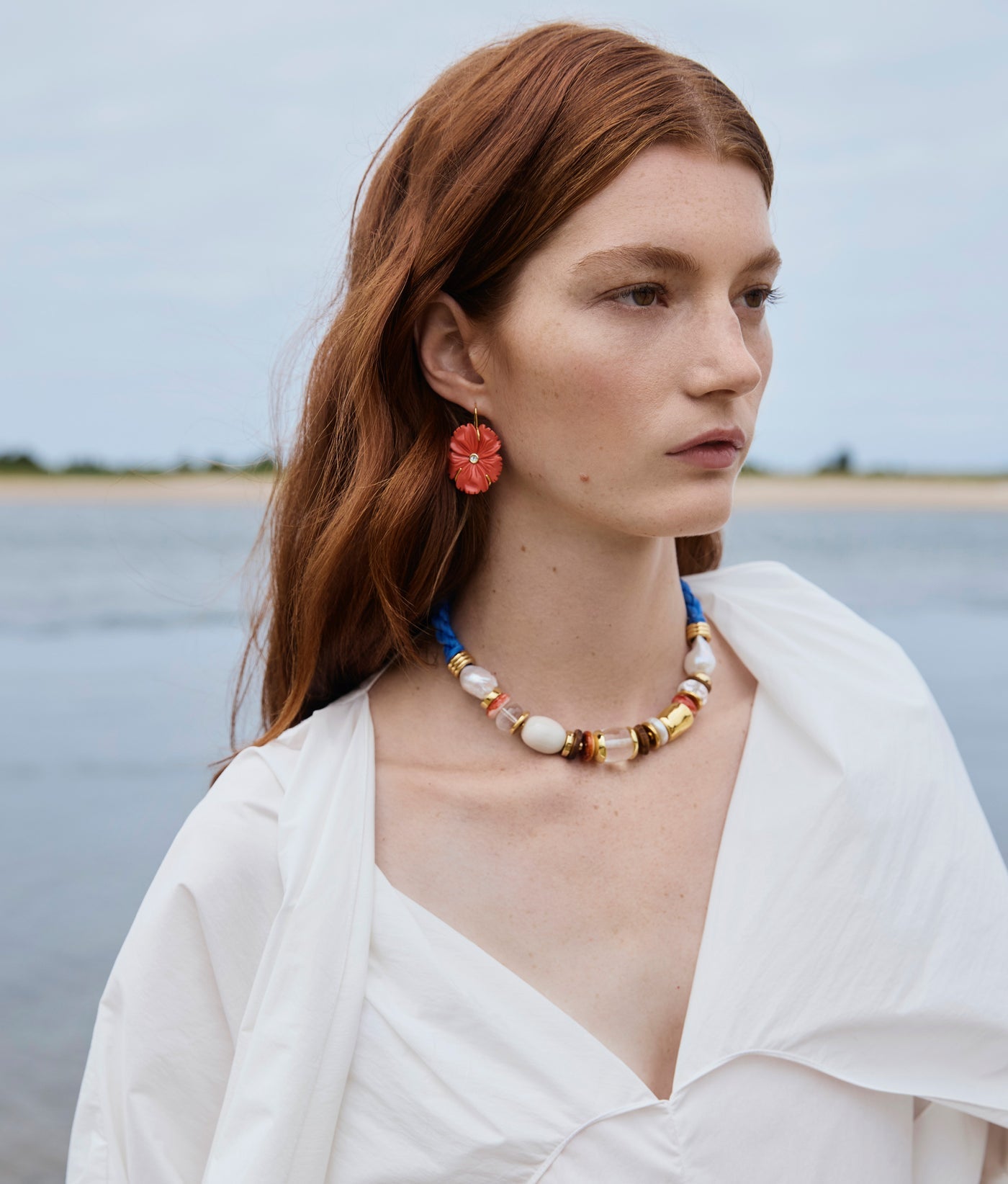 A woman with long red hair stands outdoors near the water, wearing a white blouse, a colorful beaded necklace, and Lizzie Fortunato’s New Bloom Earrings.
