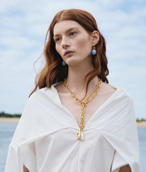 A woman with long red hair wears a white top and the Lizzie Fortunato Spiral Shell Necklace, standing outdoors against a blurred backdrop of sky and water.
