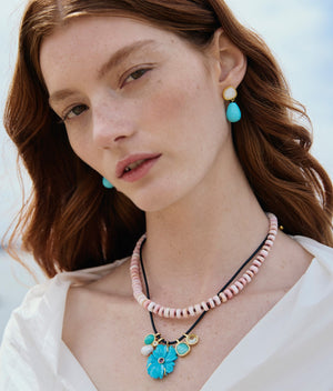 A woman with fair skin and red hair wears blue and gold earrings, a white blouse, and the Lizzie Fortunato Shibori Flower Necklace—a black leather cord layered with pink and black-beaded strands—her expression neutral.