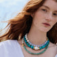 A person with long red hair wears the Lizzie Fortunato Sky Stone Necklace—layered turquoise beads, baroque freshwater pearls, and green strands with gold-plated accents—over a white top, standing outdoors on a sunny day.
