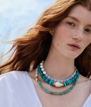 A person with long red hair wears the Lizzie Fortunato Sky Stone Necklace—layered turquoise beads, baroque freshwater pearls, and green strands with gold-plated accents—over a white top, standing outdoors on a sunny day.