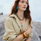 Woman with long red hair wearing a beige shirt, the Lizzie Fortunato Blue Pond Necklace, blue earrings, amber bangles, and a brown belt, standing outdoors near rocks.
