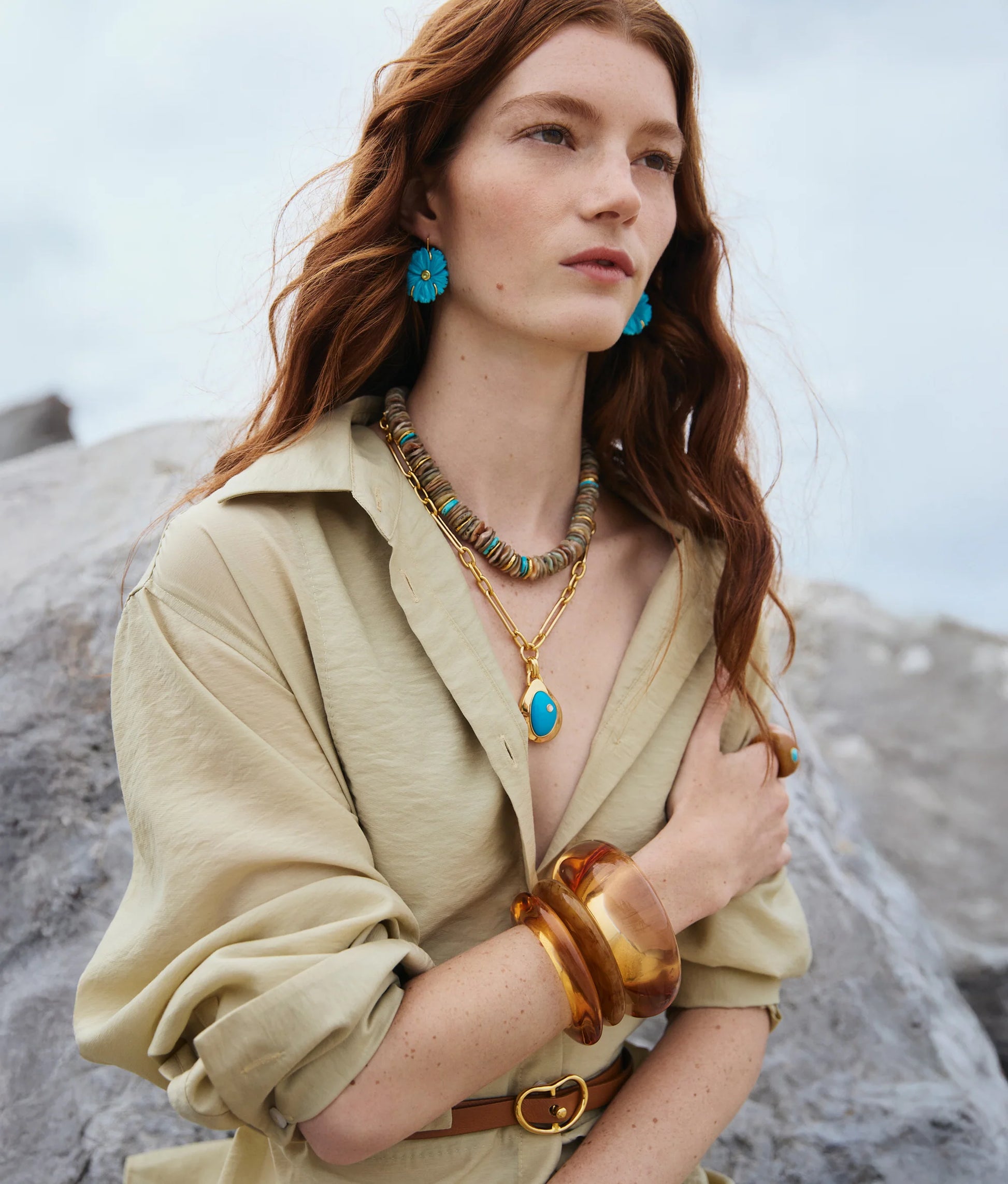 Woman with long red hair wearing a beige shirt, the Lizzie Fortunato Blue Pond Necklace, blue earrings, amber bangles, and a brown belt, standing outdoors near rocks.