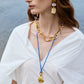 Woman wearing a white blouse, chunky gold necklace, Lizzie Fortunato Blue Point Necklace with a gold-plated oyster pendant, and gold and white statement earrings, standing near water.