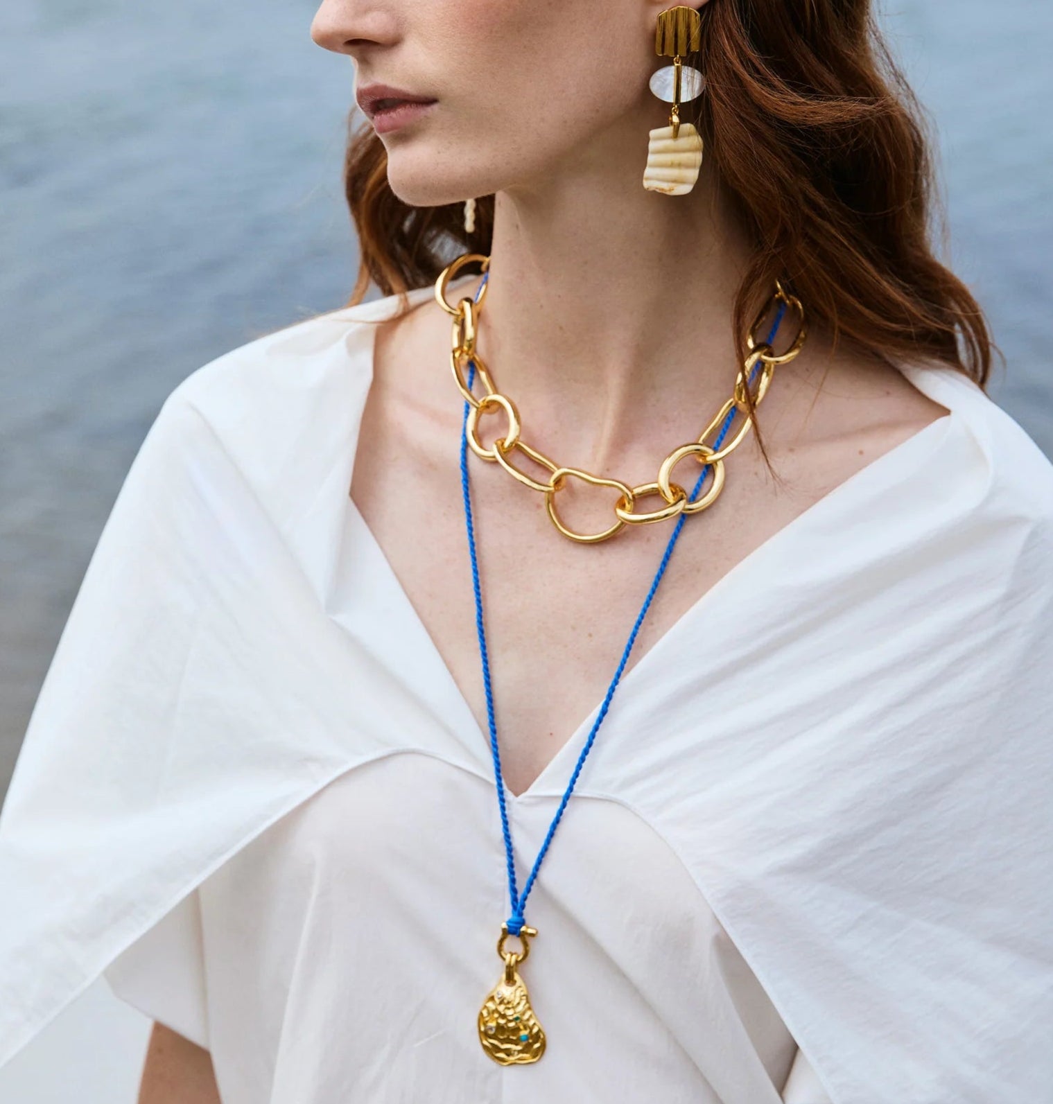 Woman wearing a white blouse, chunky gold necklace, Lizzie Fortunato Blue Point Necklace with a gold-plated oyster pendant, and gold and white statement earrings, standing near water.