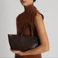 Wearing a sleeveless brown turtleneck, someone holds Mansur Gavriel's Mini Everyday handbag in dark brown Italian spazzolato leather featuring an adjustable crossbody strap against a plain background.