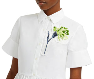 The person is wearing Erdem's Short Sleeve Embroidered Shirt Dress—a white collared shirt dress with green and white embroidered carnations on the chest, adding elegance.