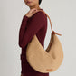 A person in a burgundy outfit holds the Mansur Gavriel Fortuna Raffia Bag, featuring a woven beige texture with a knotted leather strap.