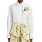 A person is wearing an Erdem Long Sleeve Embroidered Shirt, oversized and in white cotton poplin with a floral design on the left, paired with a beige skirt featuring green embroidered floral patterns.