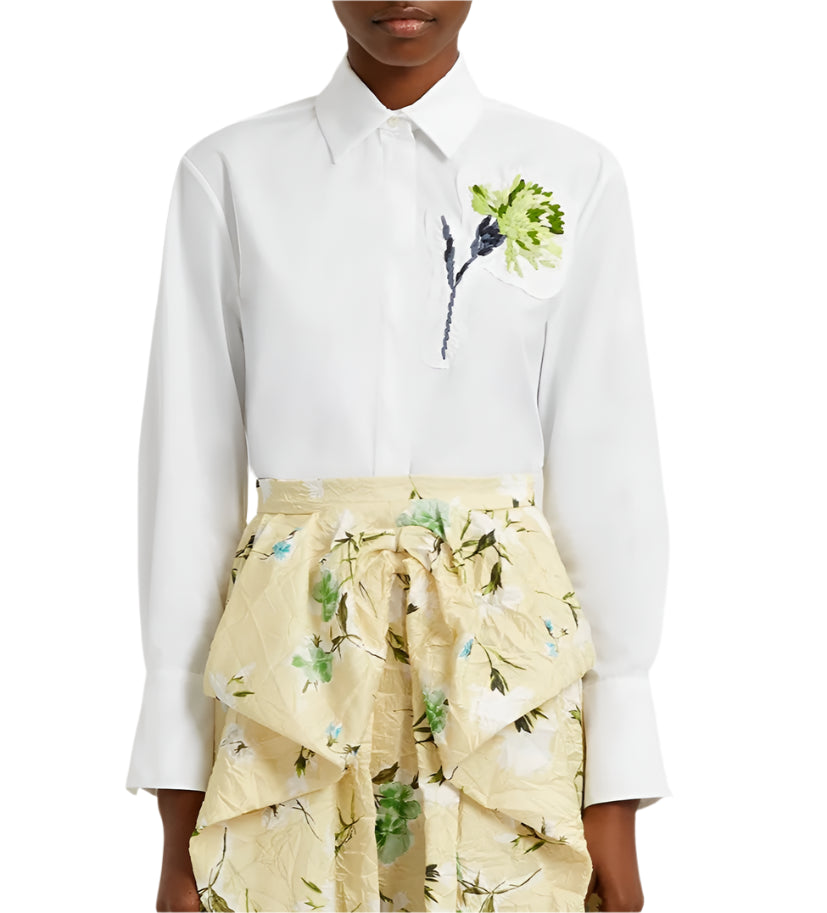 A person is wearing an Erdem Long Sleeve Embroidered Shirt, oversized and in white cotton poplin with a floral design on the left, paired with a beige skirt featuring green embroidered floral patterns.