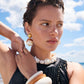 A woman in a black sleeveless top and oversized shell jewelry stands on a beach under partly cloudy skies, wearing the Lizzie Fortunato Ridge Cuff.