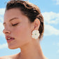 A person with short wet hair wears Lizzie Fortunato’s New Bloom Studs—large mother-of-pearl floral earrings with a turquoise center—while standing against a blue sky with scattered clouds.