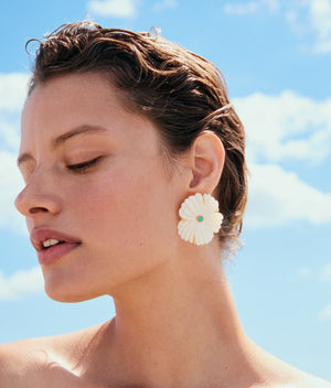 A person with short wet hair wears Lizzie Fortunato’s New Bloom Studs—large mother-of-pearl floral earrings with a turquoise center—while standing against a blue sky with scattered clouds.