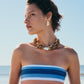 A woman in a striped, strapless top and chunky beaded jewelry stands on a beach, her look highlighted by Lizzie Fortunato's Isola Stud Earrings, gazing to the side under a clear blue sky.