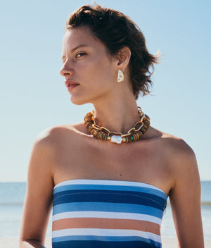 A woman in a striped, strapless top and chunky beaded jewelry stands on a beach, her look highlighted by Lizzie Fortunato's Isola Stud Earrings, gazing to the side under a clear blue sky.