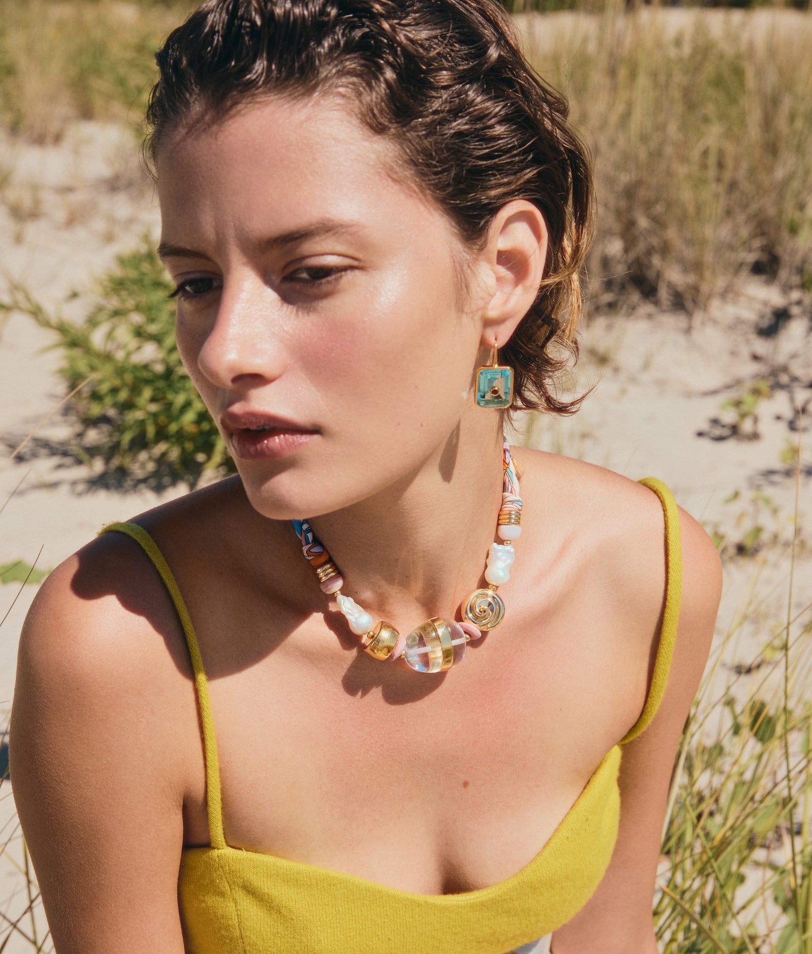 A woman with short brown hair wears a yellow spaghetti strap top, a colorful beaded necklace, and Lizzie Fortunato Tile Earrings with aqua blue glass stones while sitting outdoors on the grass.