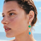 Close-up of a woman wearing Lizzie Fortunato Cala Drop Earrings—statement earrings crafted from multicolored stones and gold-plated brass—standing outdoors with a blurred beach and sky in the background.