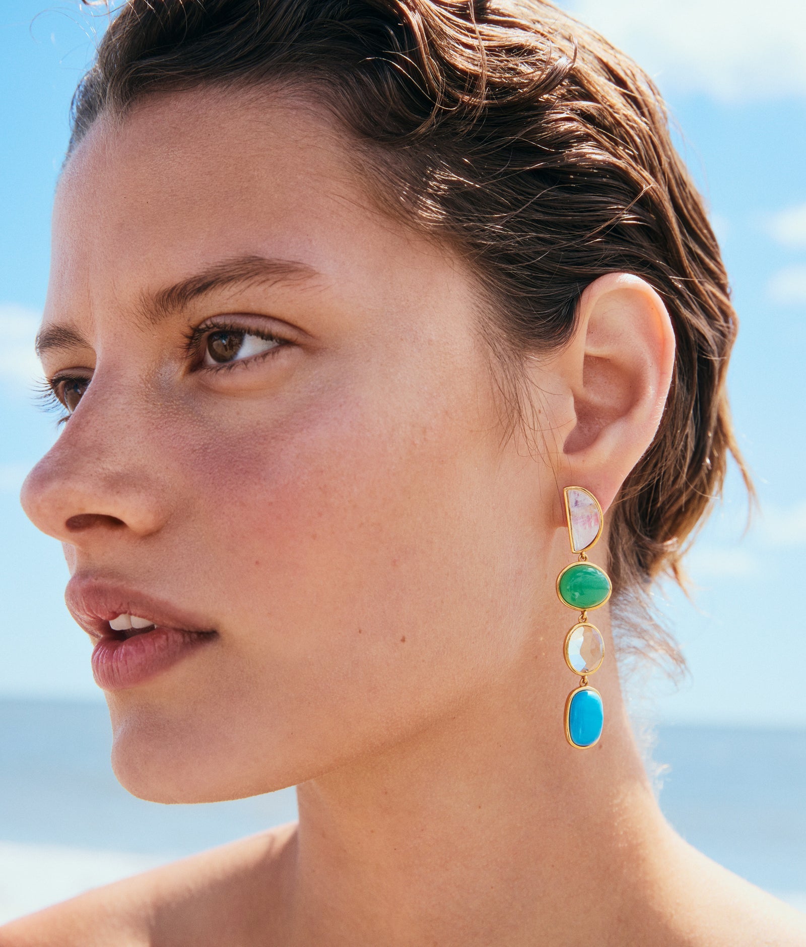 Close-up of a woman wearing Lizzie Fortunato Cala Drop Earrings—statement earrings crafted from multicolored stones and gold-plated brass—standing outdoors with a blurred beach and sky in the background.
