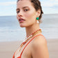A woman in an orange dress stands on a beach, wearing Lizzie Fortunato’s Marina Drop Earrings. Layered pearl necklaces and the tranquil ocean and sky complete her summery look.