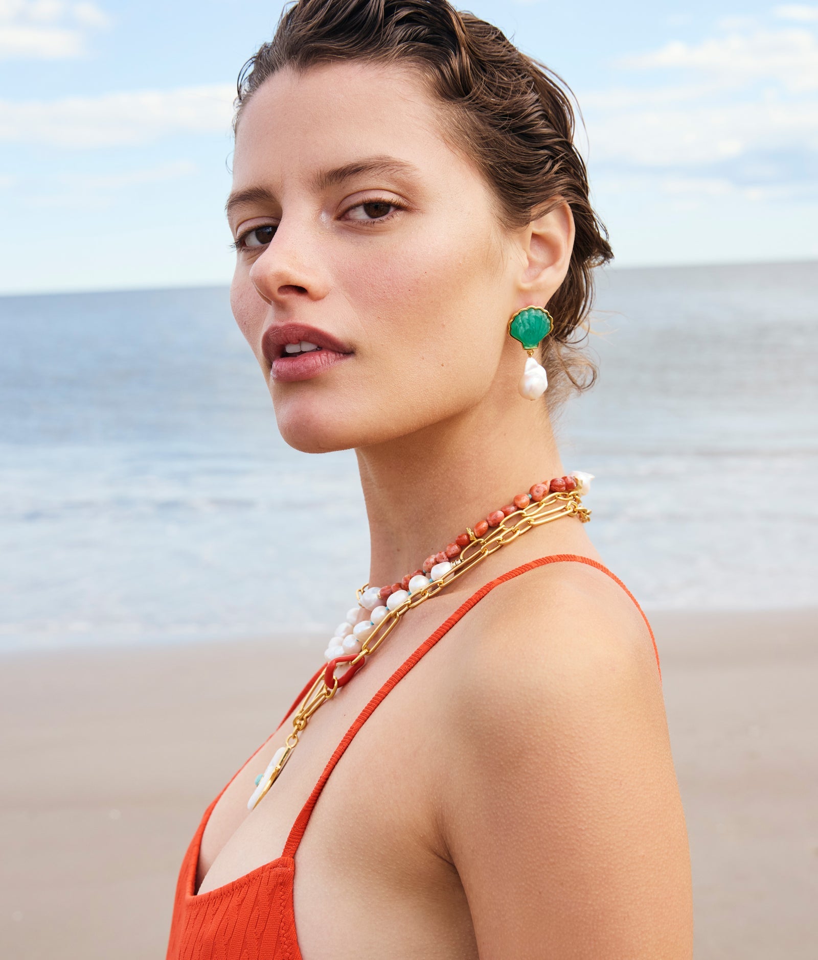 A woman in an orange dress stands on a beach, wearing Lizzie Fortunato’s Marina Drop Earrings. Layered pearl necklaces and the tranquil ocean and sky complete her summery look.
