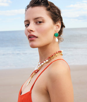 A woman in an orange dress stands on a beach, wearing Lizzie Fortunato’s Marina Drop Earrings. Layered pearl necklaces and the tranquil ocean and sky complete her summery look.