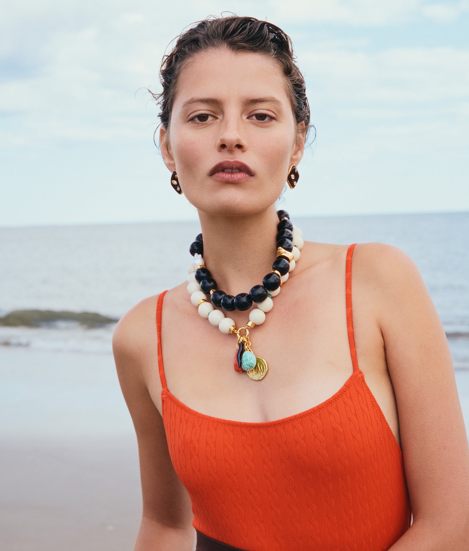 A woman stands on the beach in an orange textured swimsuit, accessorized with layered black and white bead necklaces and Lizzie Fortunato's Nico Dot Earrings, framed by the sea and sky.