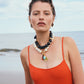 A woman in an orange sleeveless top stands on a beach, wearing the Lizzie Fortunato Oria Necklace—layered black horn beads and freshwater pearls—with gold earrings, set against a stunning sea and sky backdrop.