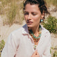 A person with short wavy hair wears a white shirt and statement jewelry, including the Lizzie Fortunato Cora Drop Earrings and a chunky gold-plated brass and amazonite beads necklace, outdoors in a sandy, grassy area.