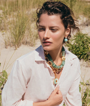 A person with short wavy hair wears a white shirt and statement jewelry, including the Lizzie Fortunato Cora Drop Earrings and a chunky gold-plated brass and amazonite beads necklace, outdoors in a sandy, grassy area.