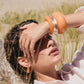 A woman in a light shirt reclines outdoors, shielding her eyes with her hand. She wears chunky orange and clear bracelets and Lizzie Fortunato Organic Mini Gold Hoops. Grassy sand appears in the background.
