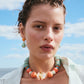 A woman with short, wavy hair wears Lizzie Fortunato Citrus Splash Earrings and a pastel beaded necklace, standing outdoors beneath a blue sky with clouds.