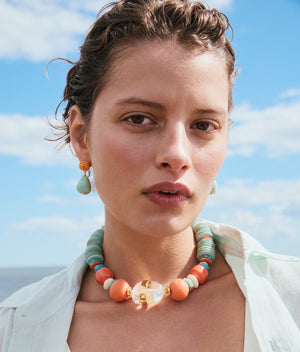 A woman with short, wavy hair wears Lizzie Fortunato Citrus Splash Earrings and a pastel beaded necklace, standing outdoors beneath a blue sky with clouds.
