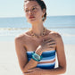 A woman stands on the beach with one hand on her chest, wearing Lizzie Fortunato Organic Gold Hoops, a striped strapless top, gold jewelry, and a large blue bracelet. The ocean waves crash in the background.