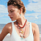 A woman in a white swimsuit and layered colorful necklaces wears Lizzie Fortunato Organic Mini Gold Hoops, standing on a beach with the ocean and blue sky behind her.