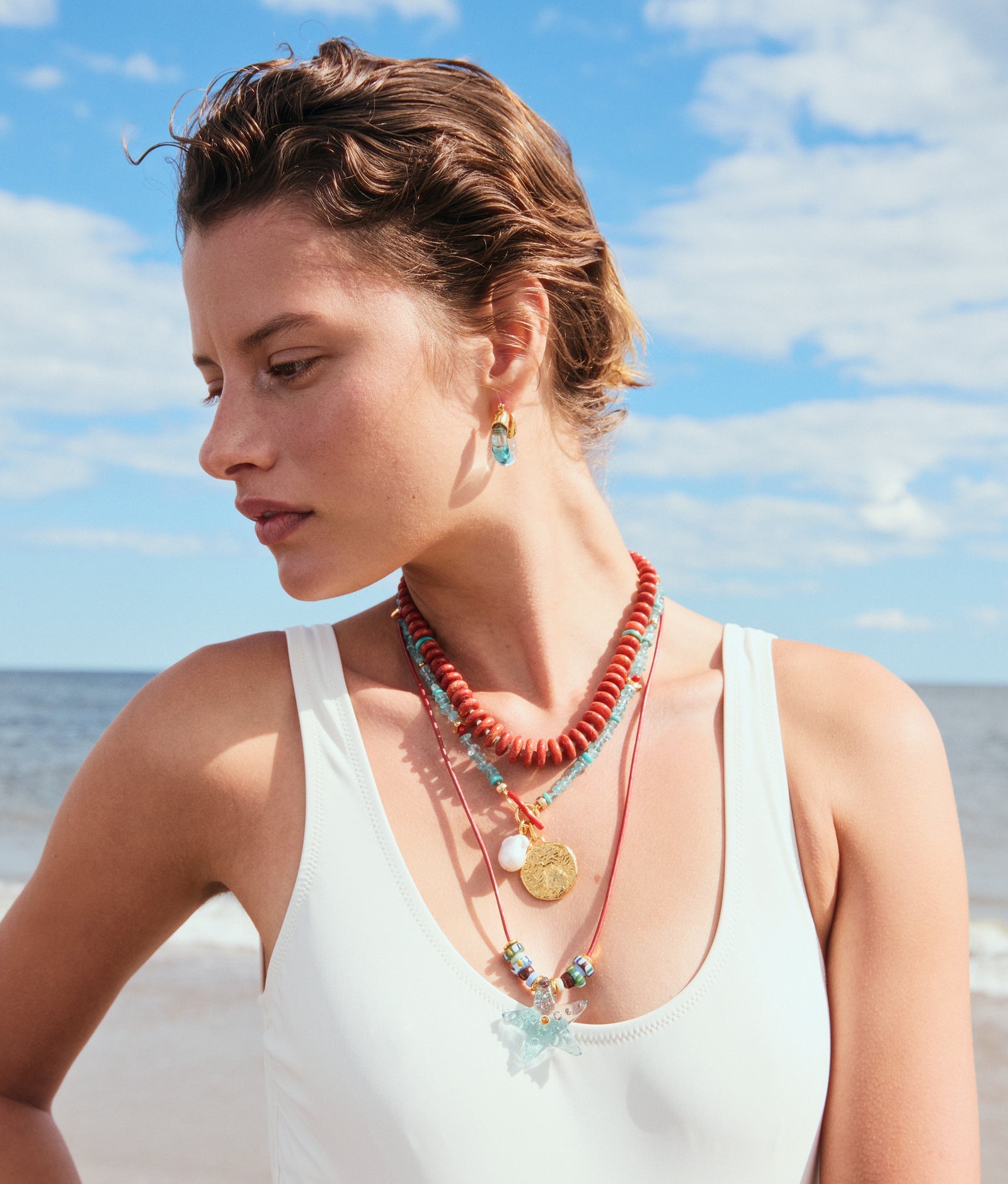 A woman in a white swimsuit and layered colorful necklaces wears Lizzie Fortunato Organic Mini Gold Hoops, standing on a beach with the ocean and blue sky behind her.