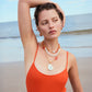 A woman in an orange tank top poses on a beach with her arm raised, wearing the Lizzie Fortunato Sirena Shell Necklace and matching earrings, with the ocean in the background.
