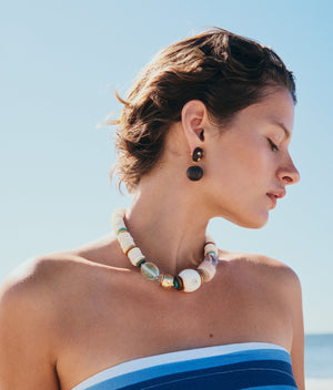 A person stands outdoors against a clear blue sky, wearing a striped blue and white top, dark round earrings, and the Coquina Necklace by Lizzie Fortunato.