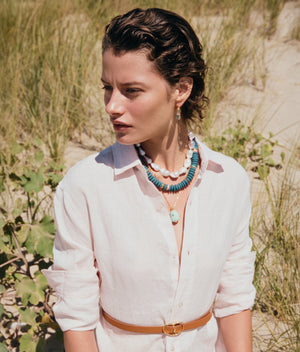 A woman with short, wet hair wearing a white button-down and brown belt sits outdoors among tall grass and sand, showcasing the Claude Necklace by Lizzie Fortunato with gold-plated brass baroque pearls.