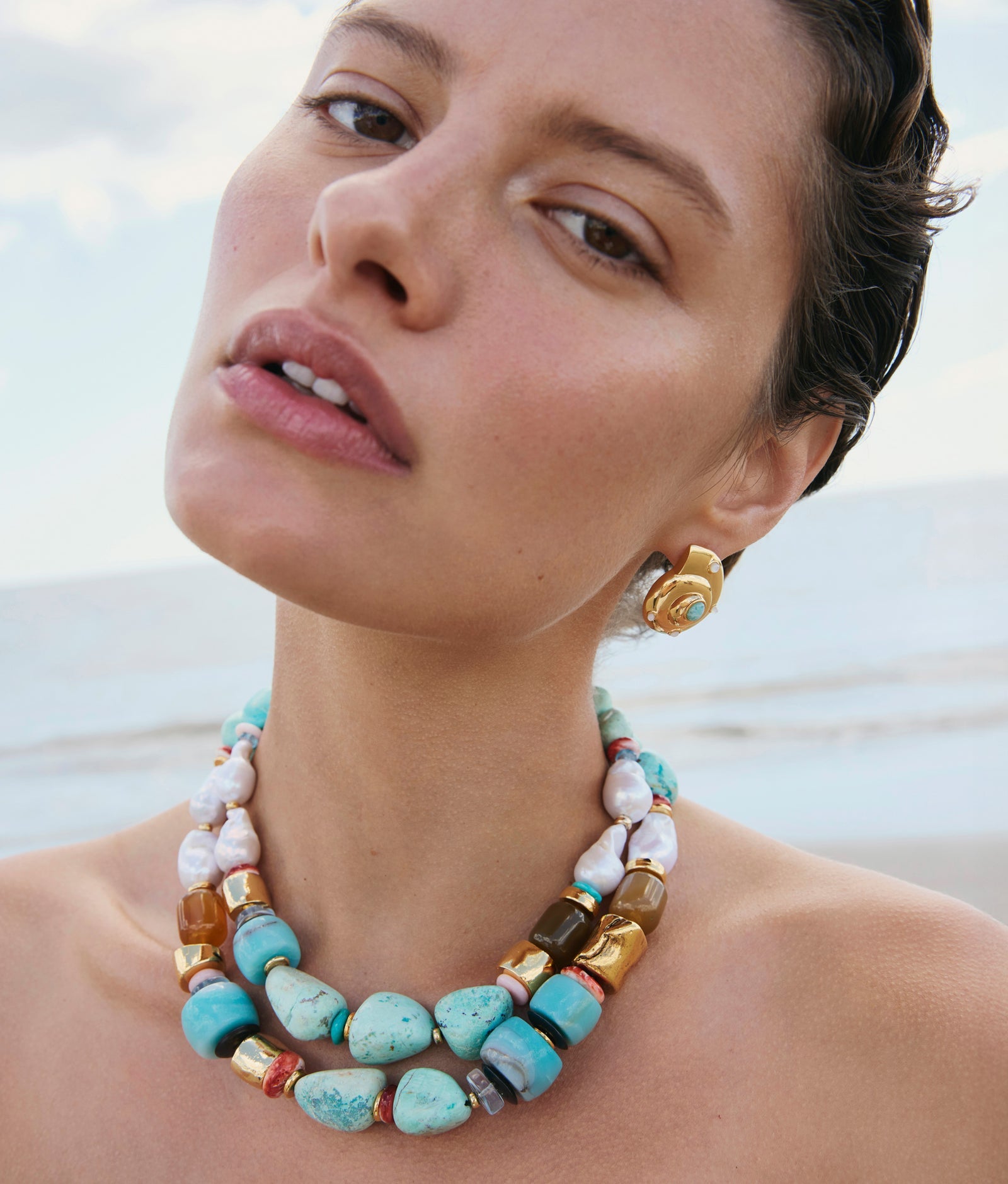 A woman wearing gold earrings and the Lizzie Fortunato Ariel Necklace—featuring double strands of turquoise, freshwater pearl, and gold beads—poses outdoors by the beach.