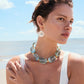 A woman in a white swimsuit stands on the beach, wearing Lizzie Fortunato's Taj Disc Earrings and a chunky translucent necklace, with the ocean and sky in the background.