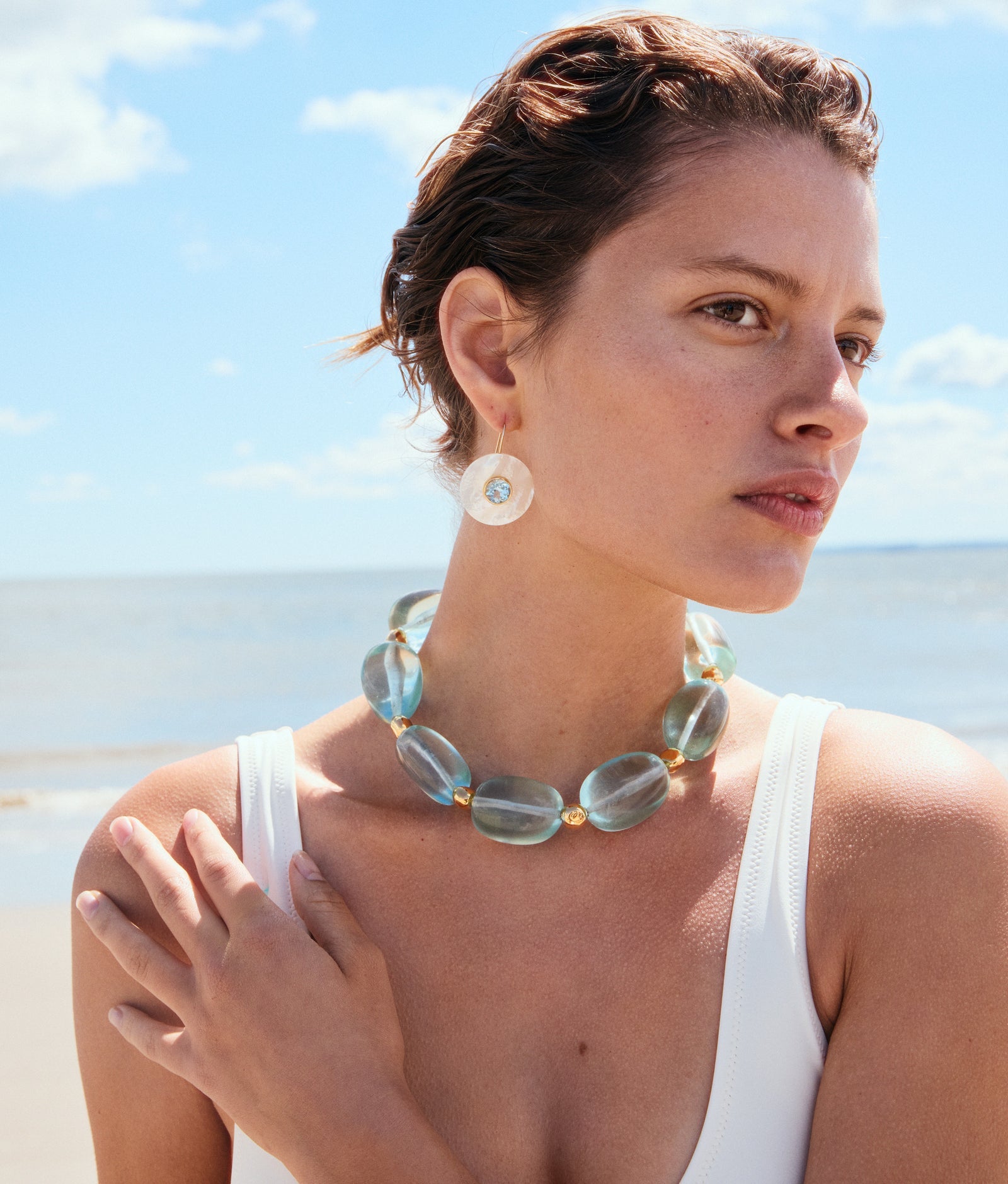 A woman in a white swimsuit stands on the beach, wearing Lizzie Fortunato's Taj Disc Earrings and a chunky translucent necklace, with the ocean and sky in the background.