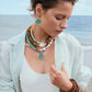 A woman wearing the Lizzie Fortunato New Bloom Necklace, chunky bracelets, a white swimsuit, a sheer light shirt, and large earrings stands by the sea, looking down.