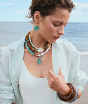 A woman wearing the Lizzie Fortunato New Bloom Necklace, chunky bracelets, a white swimsuit, a sheer light shirt, and large earrings stands by the sea, looking down.